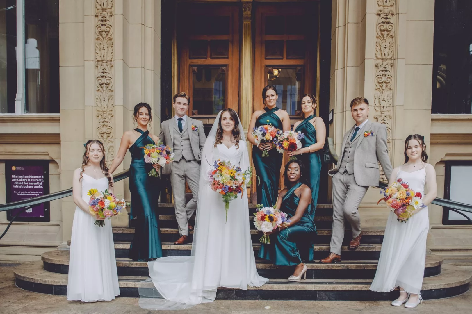 bride and her bridesmaids and groomsmen standing on the stairs holding bright and colourful bouquets outside Birmingham museum and art gallery
